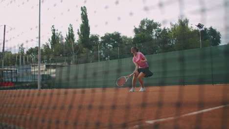 Slow-Motion-commercial-footage-of-tennis-practice-through-the-tennis-court-net.-Straight-view-of-a-female-athlete-playing-the-tennis-game.-A-teenage-sportsman-is-hitting-the-ball-during-sport-training